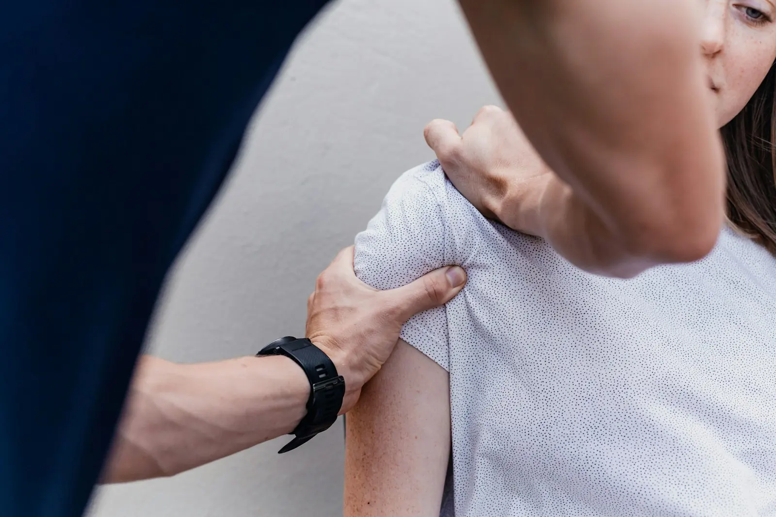 Ein Physiotherapeut im marineblauen Shirt tastet mit beiden Händen die Schulter einer Patientin ab.
